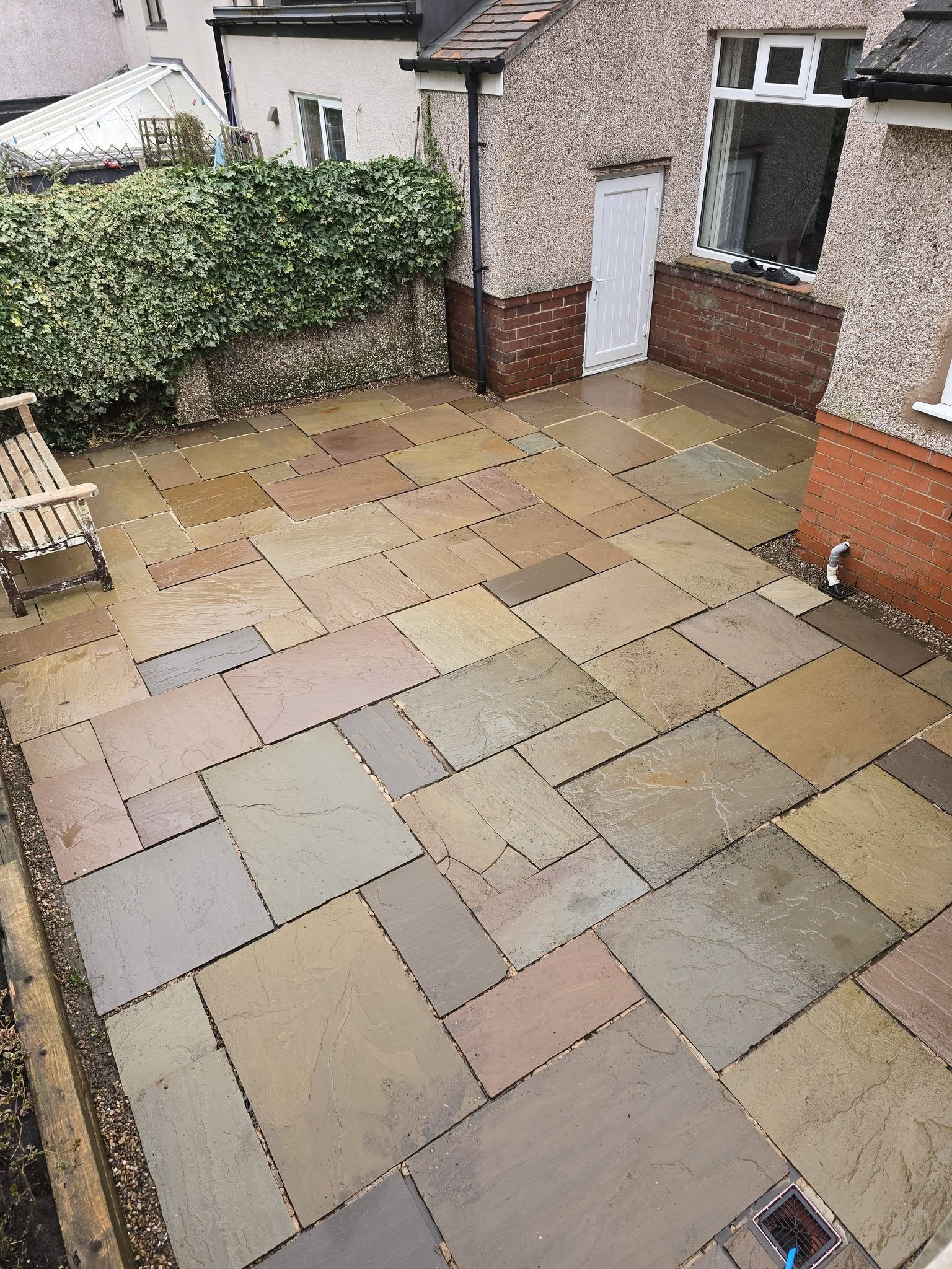 Clean, wet stone patio with a wooden bench on the left and a drain in the bottom right corner, neighboring a house with brick and stucco exterior, large window, and door.
