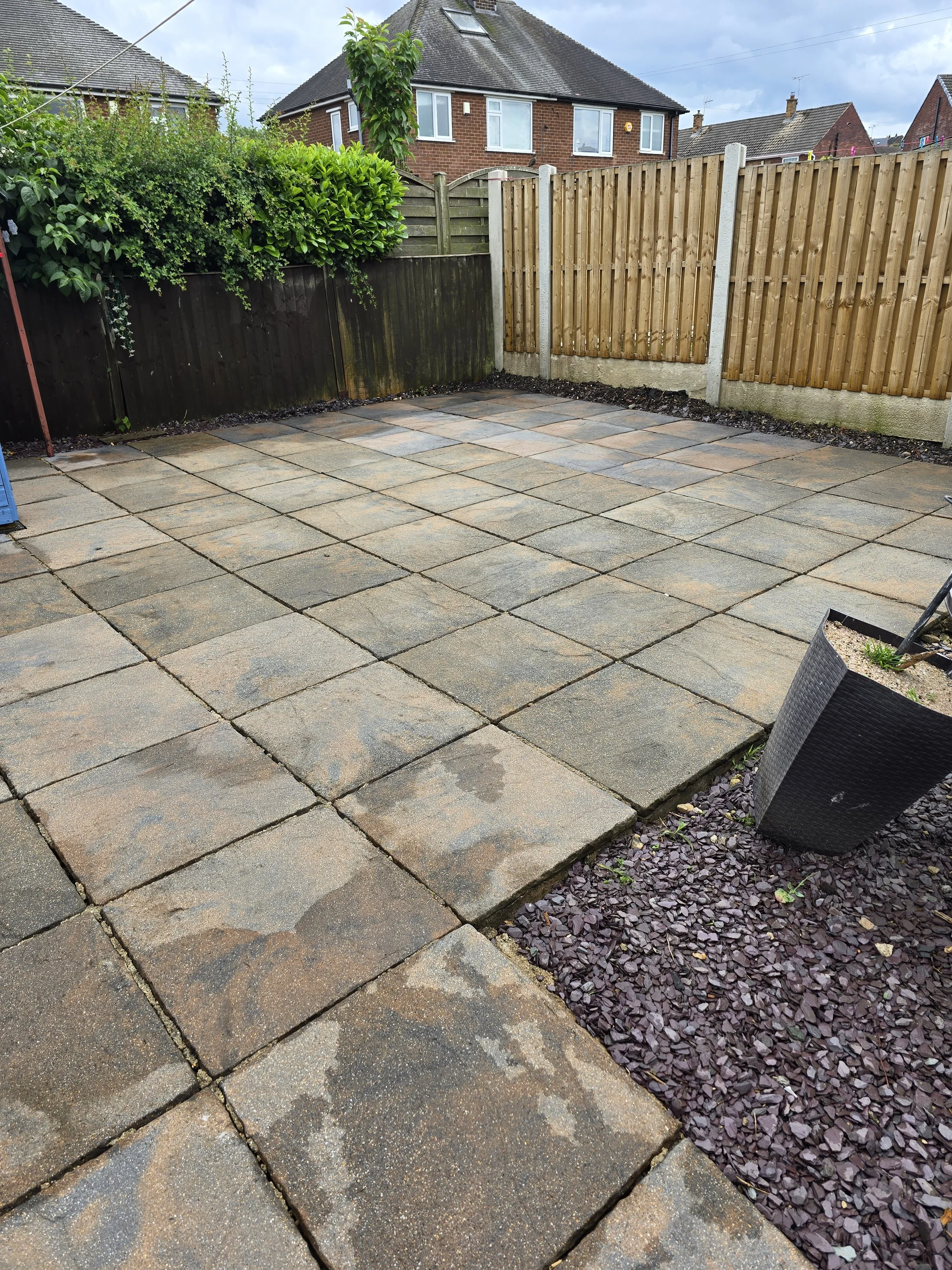 Backyard patio with stone tiles, a stone border, and a wooden fence. There are plants and a small patch of purple decorative stones at the corner of the patio, with a house and gray sky in the background.