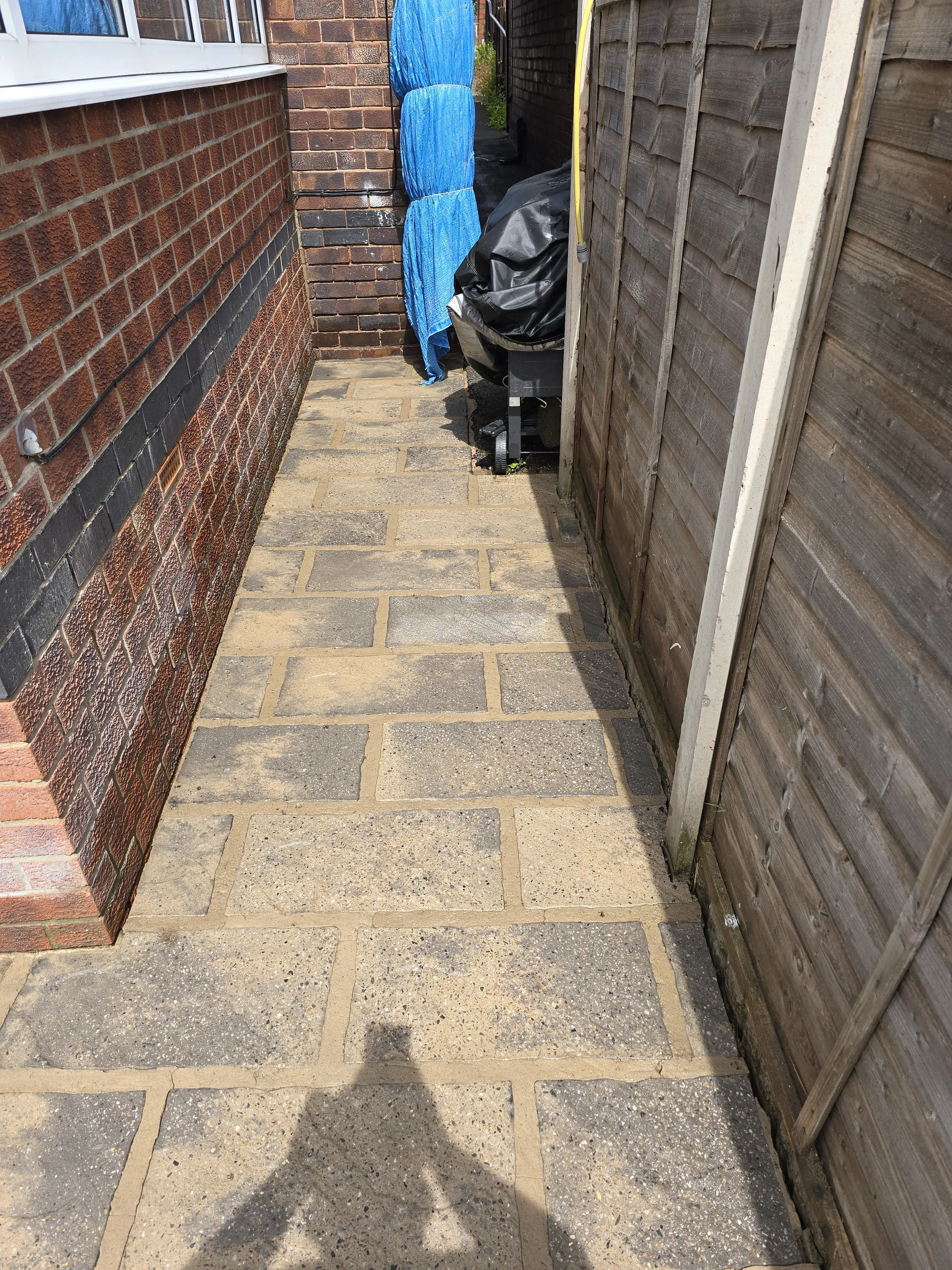 A small outdoor patio area with brick and wooden fences. The ground is paved with large concrete bricks, and there is a black barbecue grill covered with a black tarp to the side. Blue packaging or wrapping is hanging on the fence, and a yellow pole 