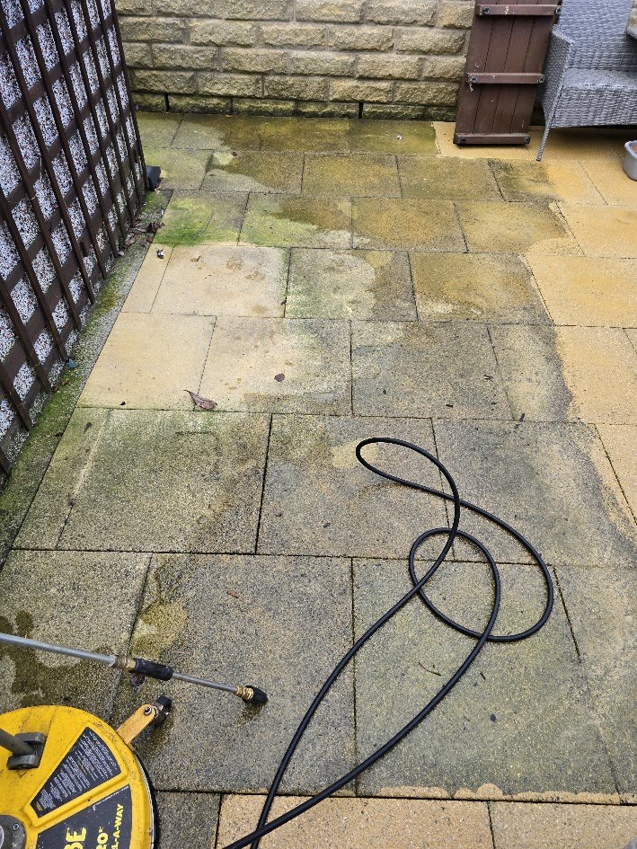 Wet and mossy outdoor patio with paving stones, garden hose, and pressure washer in the foreground; brick wall and outdoor furniture in the background.