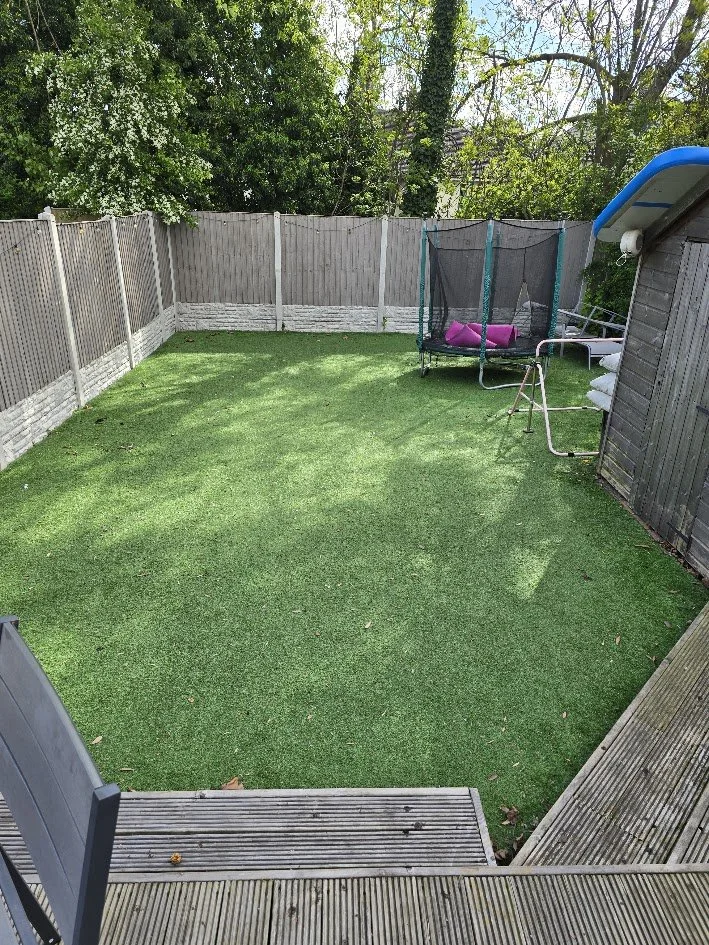 Empty backyard with artificial grass, wood fence, and a small trampoline with a purple cushion, a seating area with white chairs, and a wooden deck.