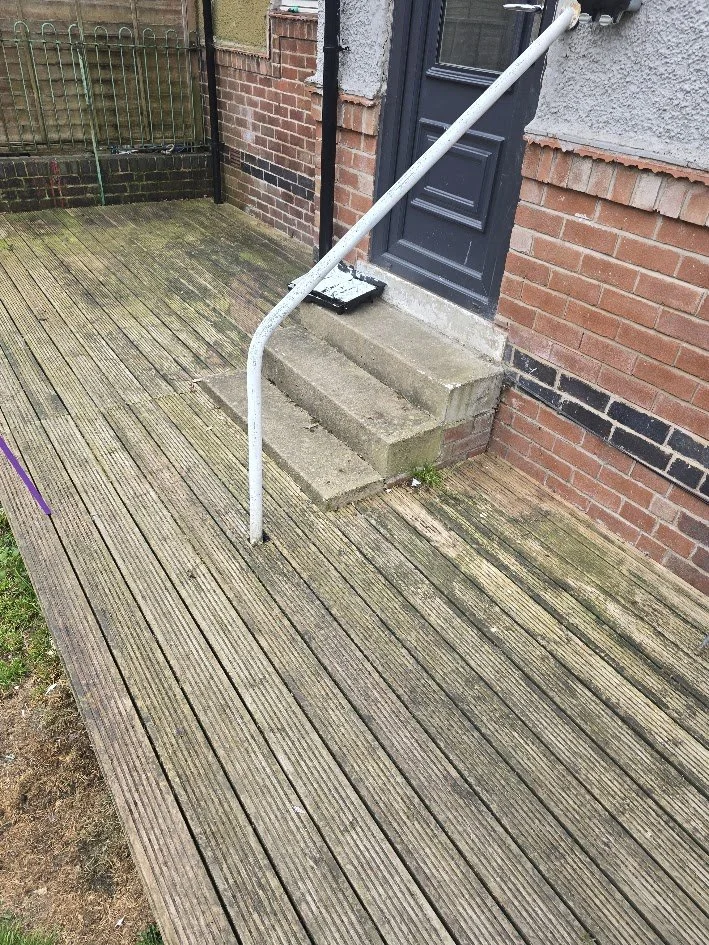 Outside view of a brick house entrance with a small wooden porch, concrete steps, a black door, and a metal hand rail.