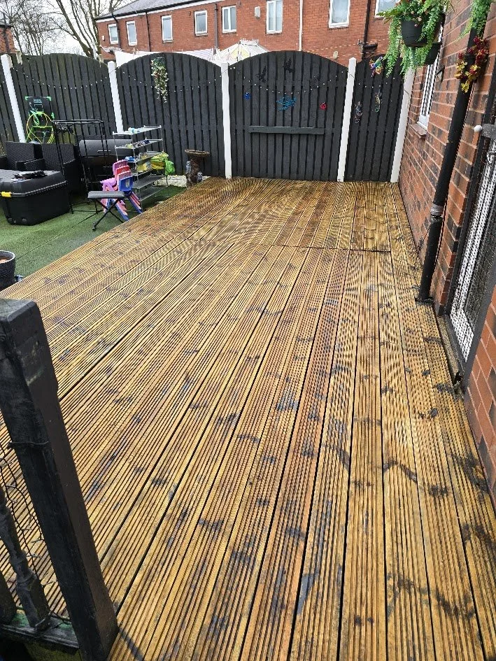 A wooden deck outdoor patio area with a black fence, potted plants, and some children's toys and storage boxes near the fence. The house wall with a window and brick exterior is visible on the right.