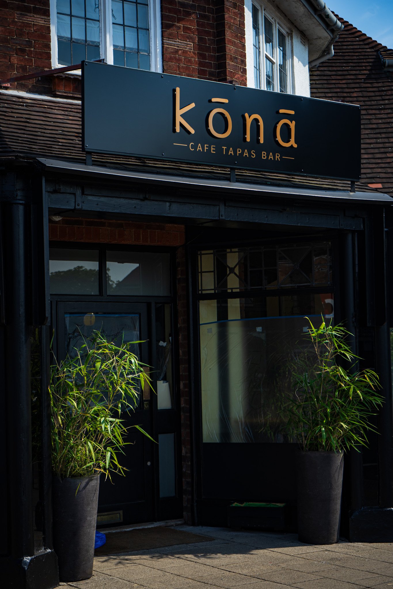 Exterior view of a restaurant called 'Kōnā Cafe Tapas Bar' with black sign, brick building, and large glass windows. Two tall potted plants are positioned on either side of the entrance, and a skyline with outdoor seating is visible through the windows.