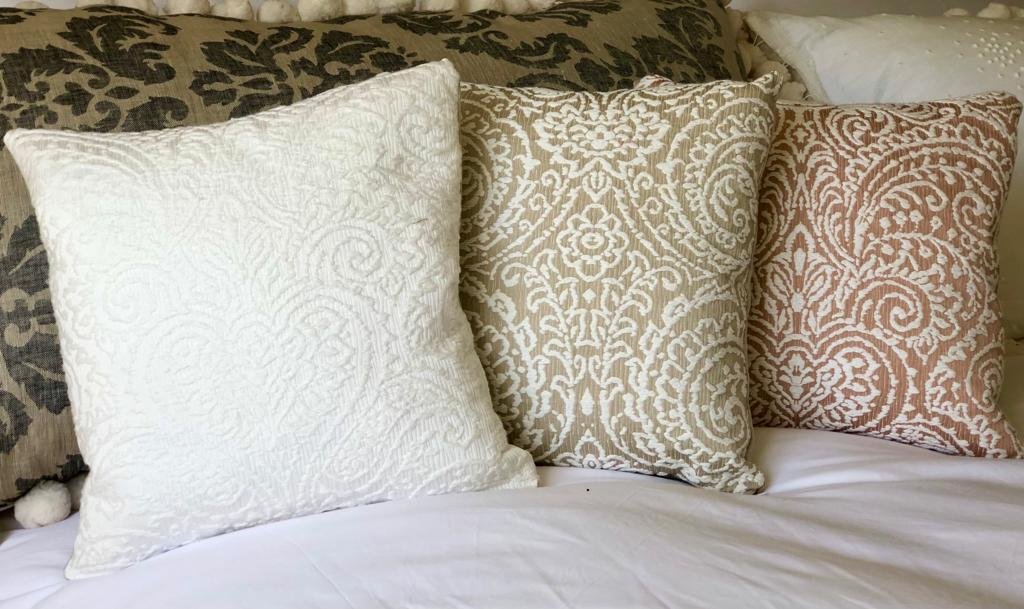 Three decorative pillows with ornate embroidered patterns placed on a bed with dark and light bedding.