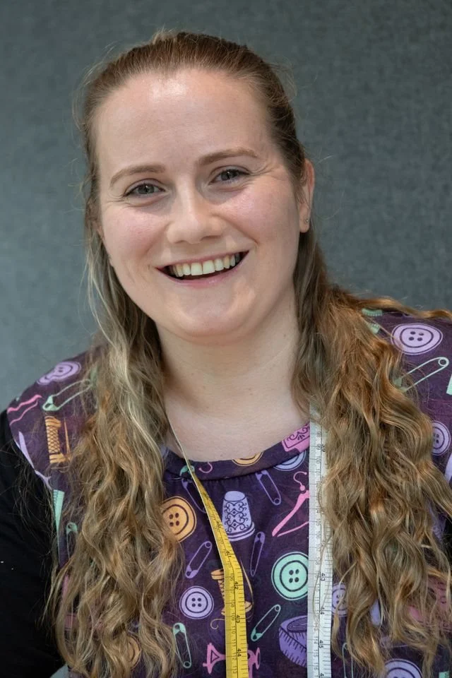 Close-up of a smiling woman with long wavy blond hair, wearing a colorful sewing-themed apron, and holding a measuring tape around her neck.