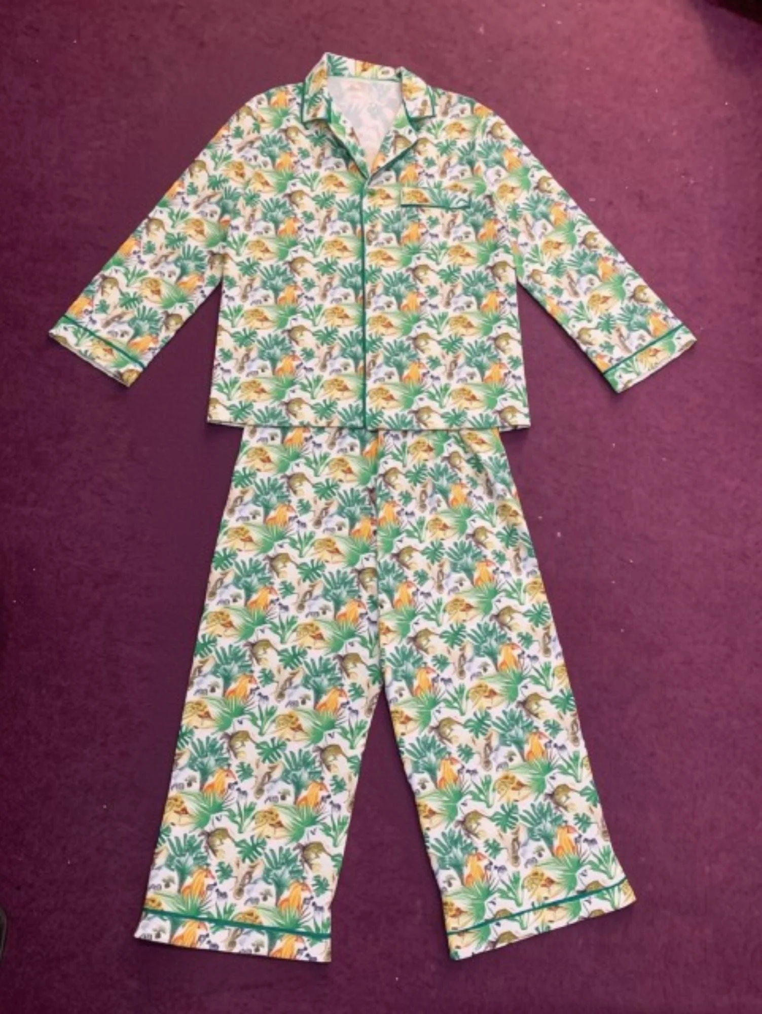 A set of colorful, tropical-themed pajamas with a button-up top and matching pants, featuring a pattern of lions, zebras, and other animals among green palm leaves on a yellow and white background, laid out on a maroon carpet.