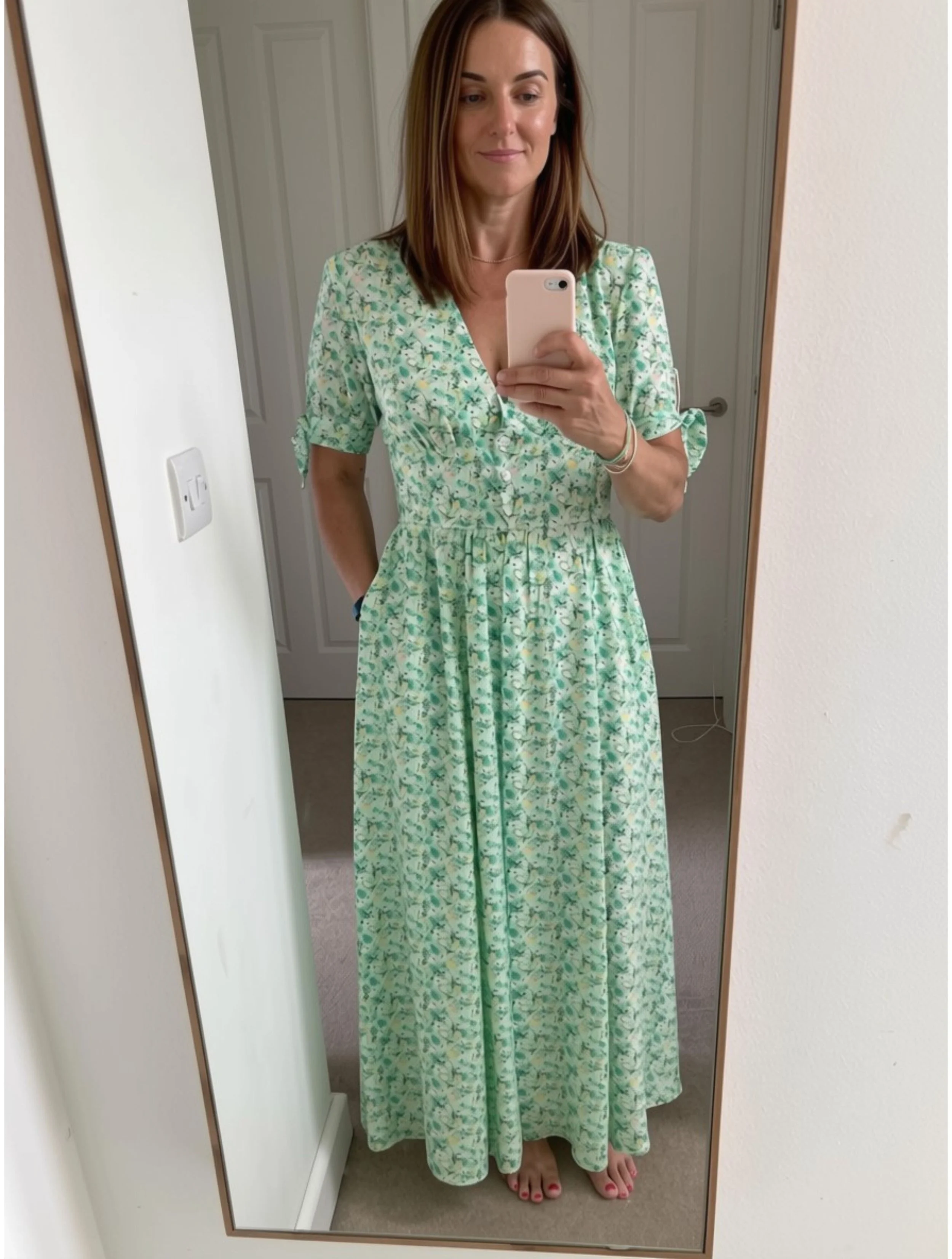 A woman wearing a green floral dress taking a mirror selfie in a room with white walls and a door in the background.