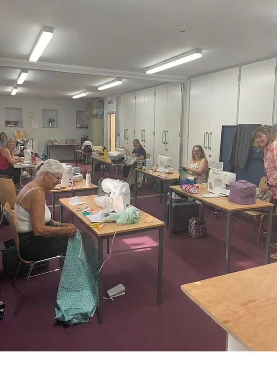 Women in a sewing class sitting at tables with sewing machines, fabric, and supplies, smiling and working.