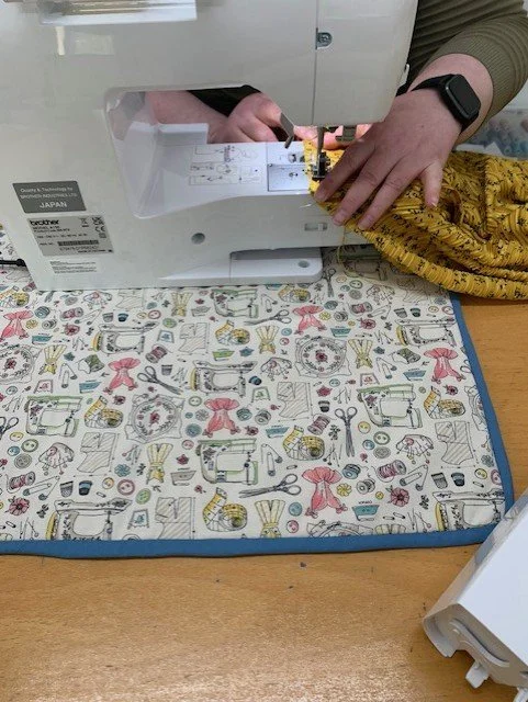 Person sewing fabric with a sewing machine on a colorful quilted tablecloth.