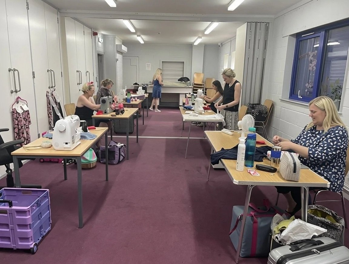 Women sitting at tables sewing fabric in a room with purple carpet, white walls, and fluorescent lighting.