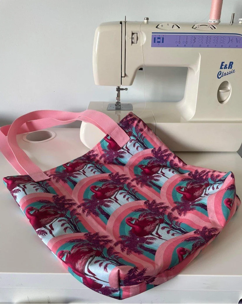 A sewing machine on a white table with a colorful tote bag with flamingos and tropical trees next to it.