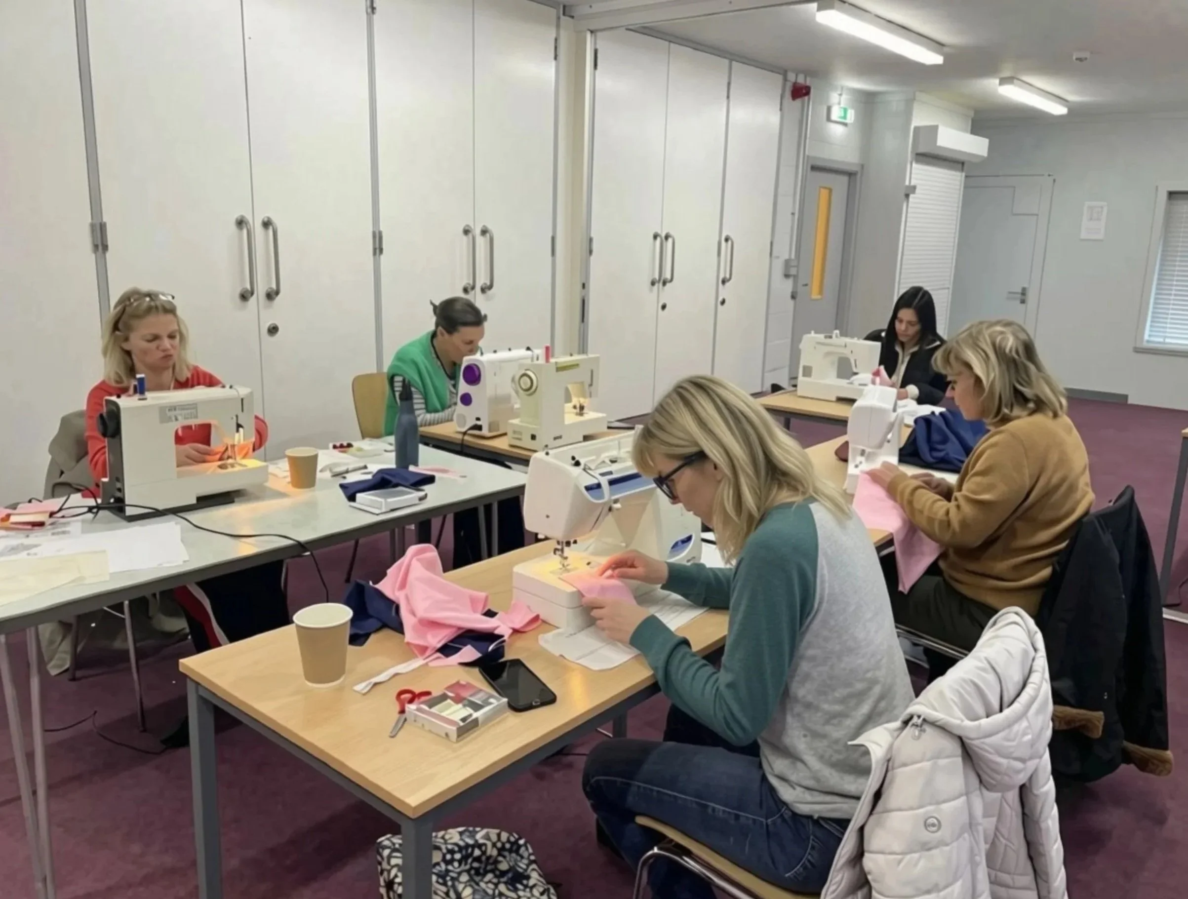 A group of women sewing in a classroom. They are seated at tables with sewing machines, working on pink and beige fabric. The room has white walls, a row of large storage cabinets, and fluorescent lighting.