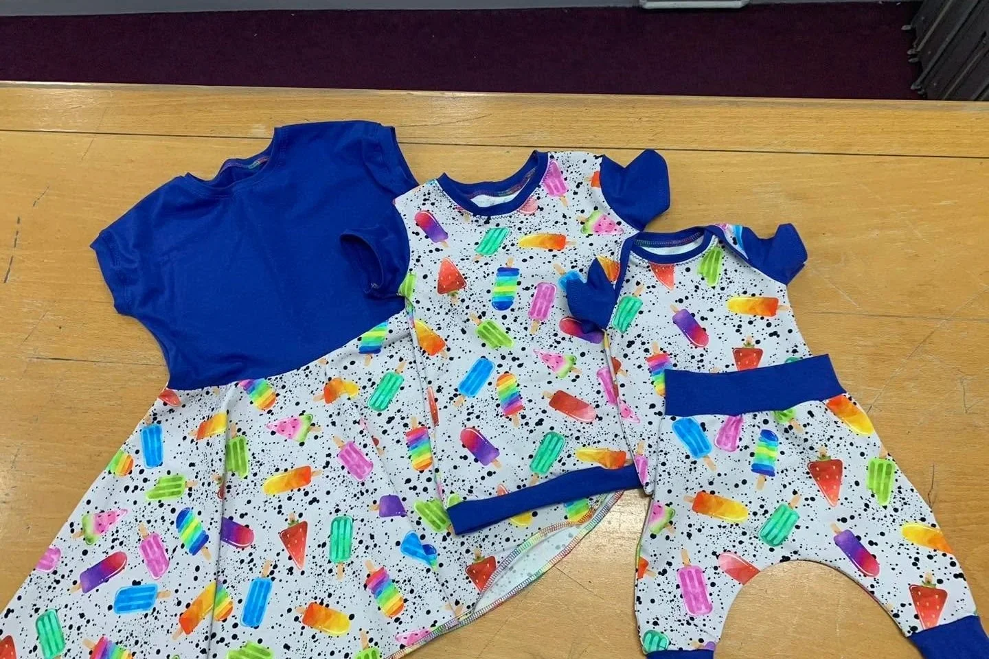 Two matching children's outfits with a colorful ice cream and popsicle pattern, featuring blue sleeves and waistband, laid out on a wooden surface.