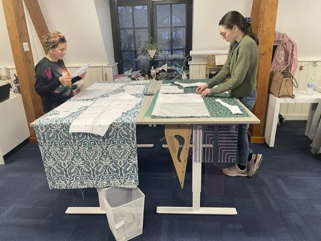 Two women working with fabric at a worktable in a craft room, with various fabric pieces and patterns spread out. One woman is reading a manual, while the other is arranging fabric on a cutting mat.