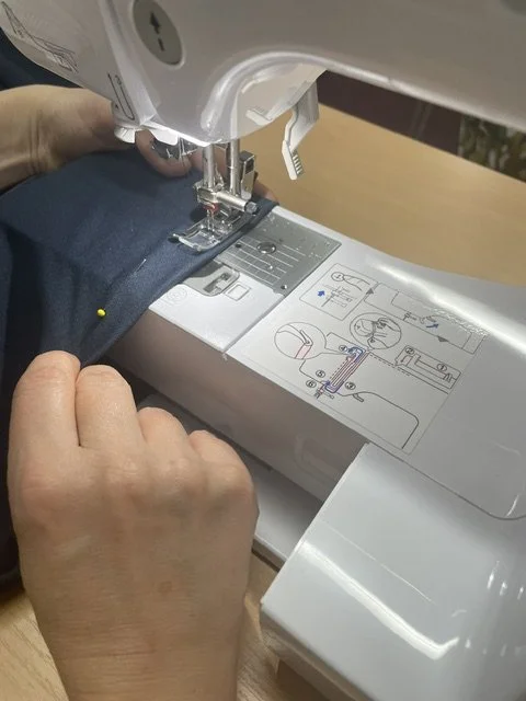 Close-up of a person sewing dark fabric on a sewing machine with instructional diagrams on the machine's surface.