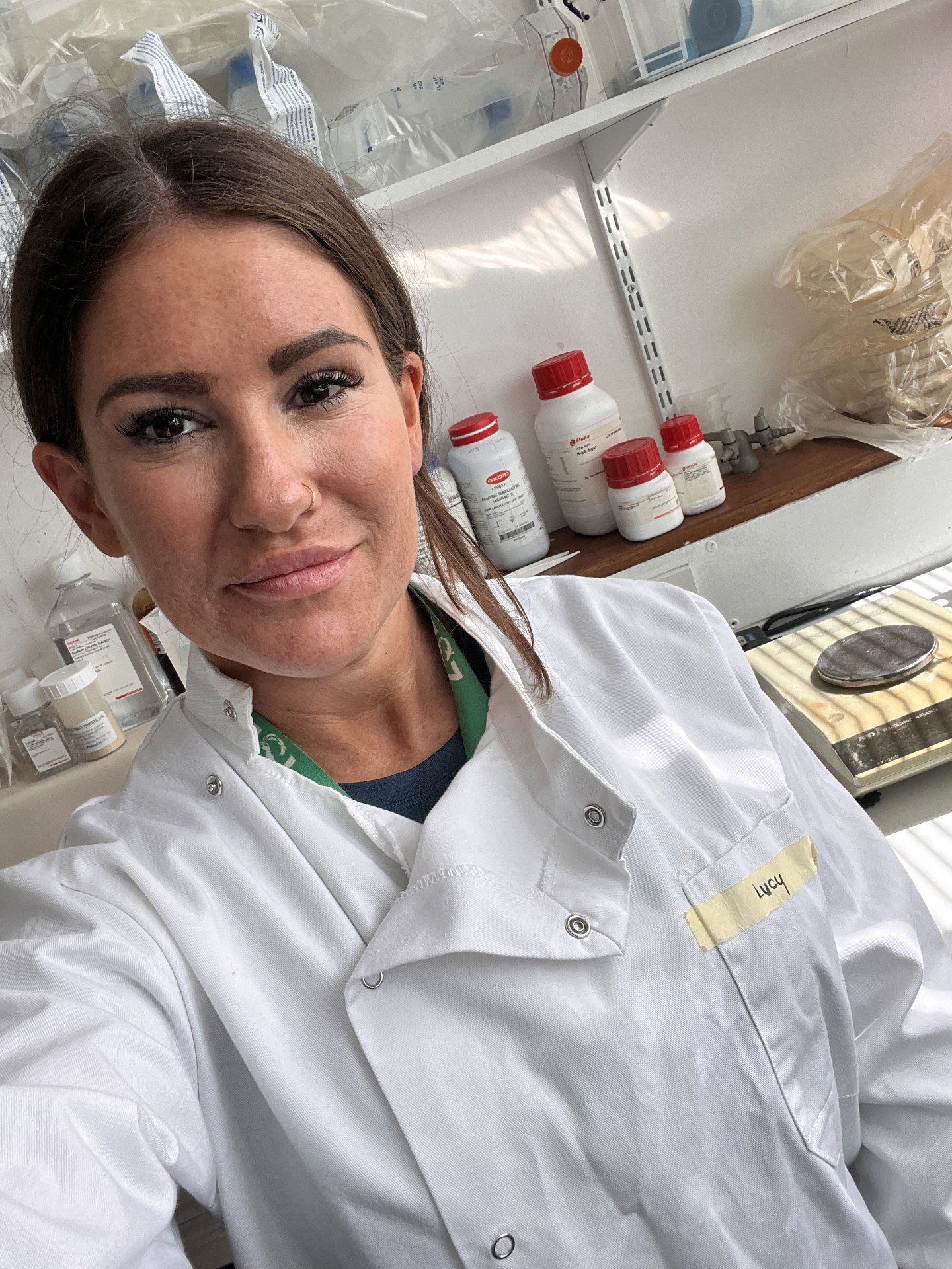 A woman in a white lab coat with a yellow name tag that reads "LUCY" is taking a selfie in a laboratory. The background shows shelves with medication bottles, medical supplies, and a scale.