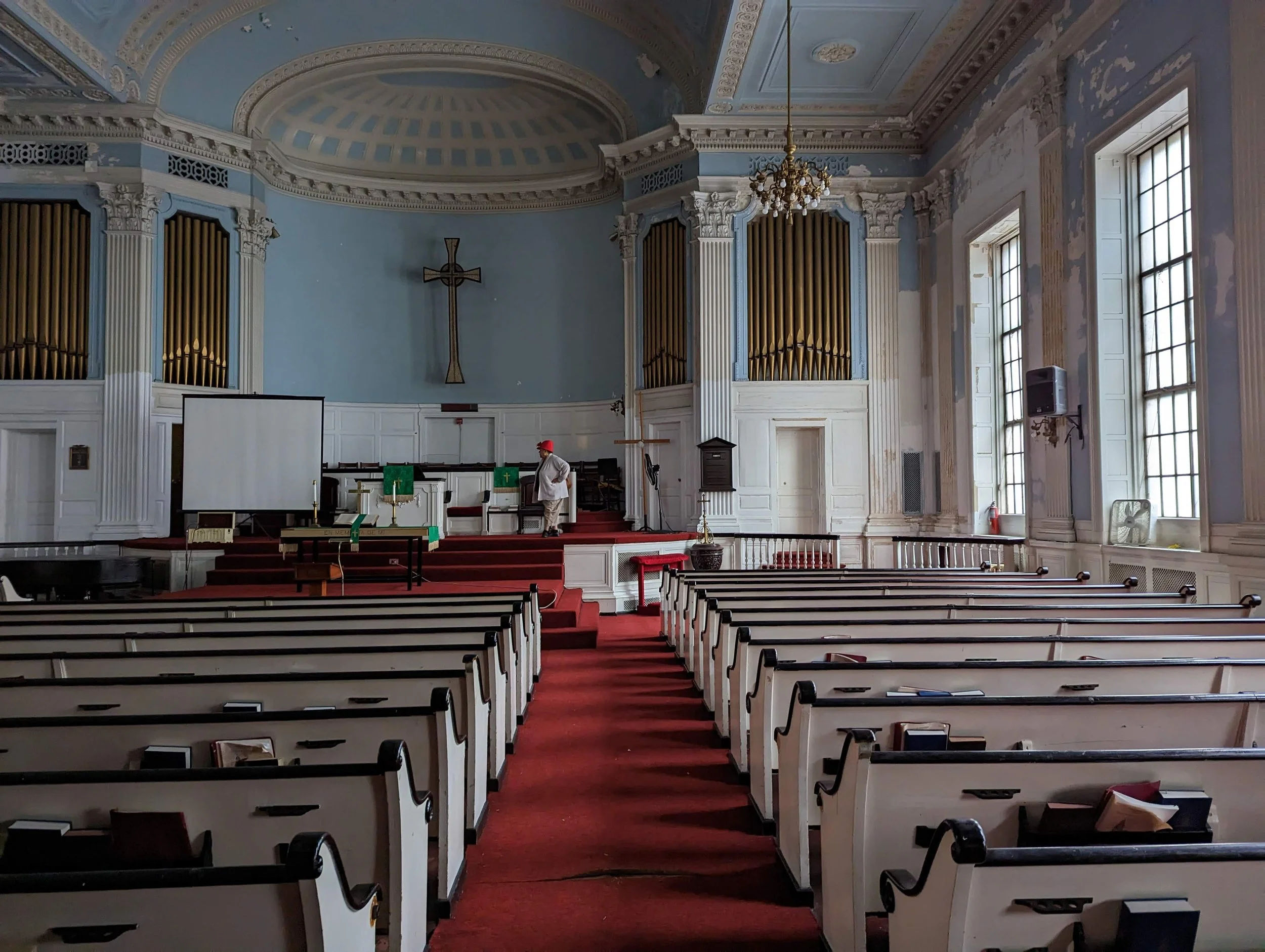 Interior of a church with white pews, a red carpet aisle, tall windows, pipe organ, chandelier, cross, and altar area. A person wearing a red hat and white shirt is at the front.