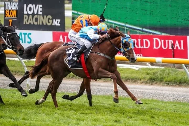 Horse race with three jockeys competing on a dirt track. The leading horse is brown with a blue and yellow bridle, followed by other horses.
