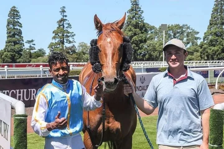 Two men standing on a racetrack with a brown racehorse, which has black protective ear covers. The man on the left is holding the horse's bridle and wearing a colorful jockey silks, while the man on the right is wearing a light blue polo shirt and cap. There are trees and race track railings in the background.