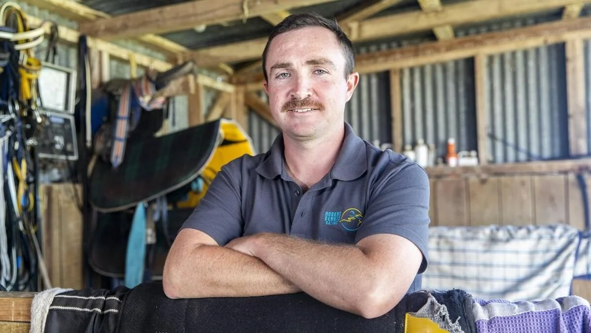 A man with short dark hair and a mustache, wearing a gray polo shirt with a logo, sitting with his arms crossed on a table inside a wooden shed with various tools and equipment in the background.