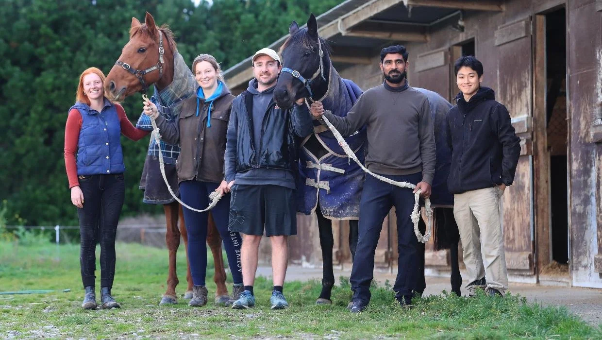 Group of six people and two horses standing outside a barn, smiling at the camera.