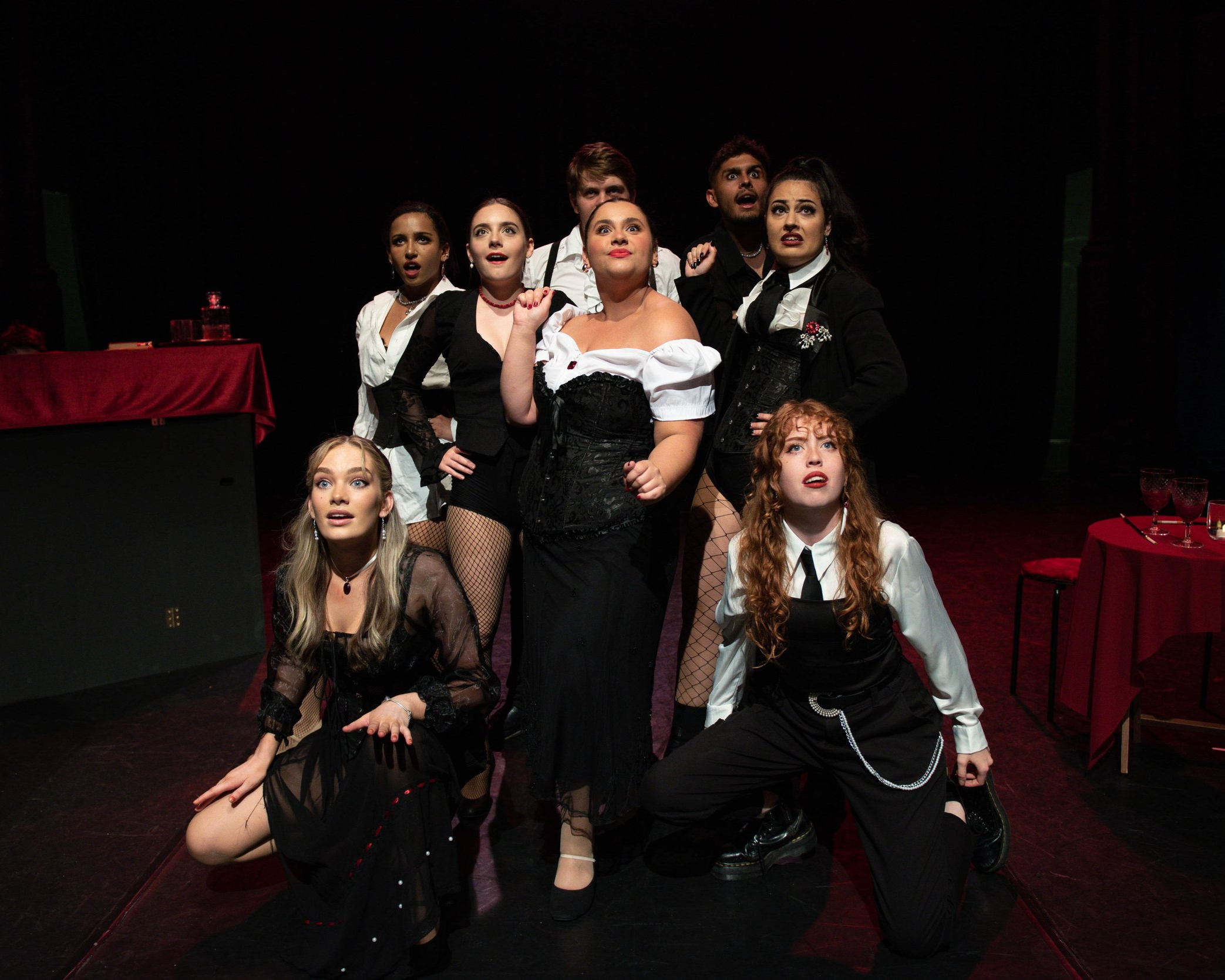 Group of six young adults, five women and one man, dressed in dark and white attire, posing dramatically in a theater setting with a black background and red tablecloths.