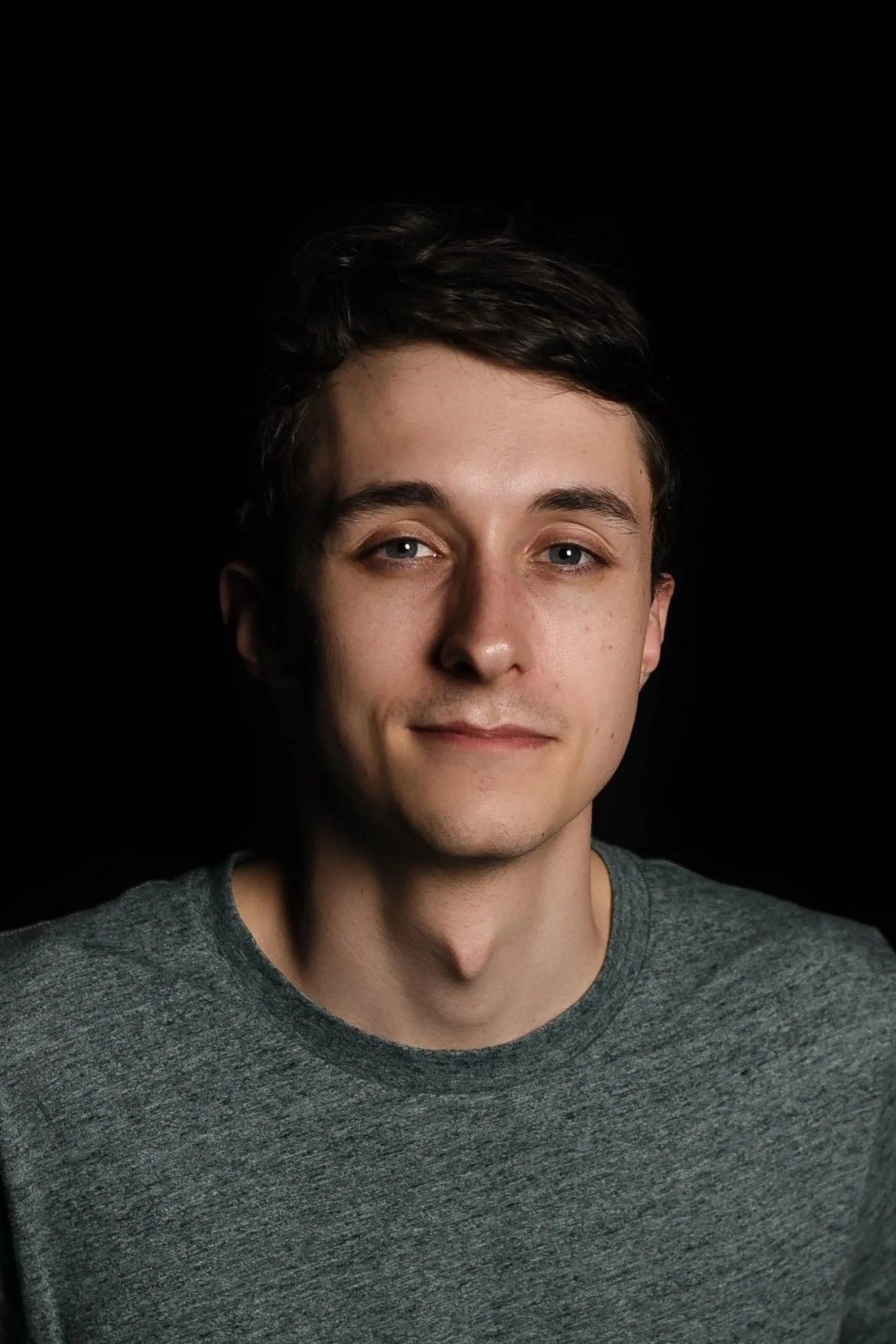 Close-up of a young man with short dark hair, light complexion, wearing a gray t-shirt, against a black background.