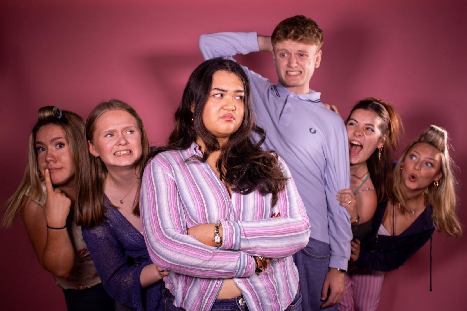 Group of six young adults, five women and one man, with varied facial expressions and poses against a pink background. Some appear confused, nervous, or surprised, while others look playful or teasing.