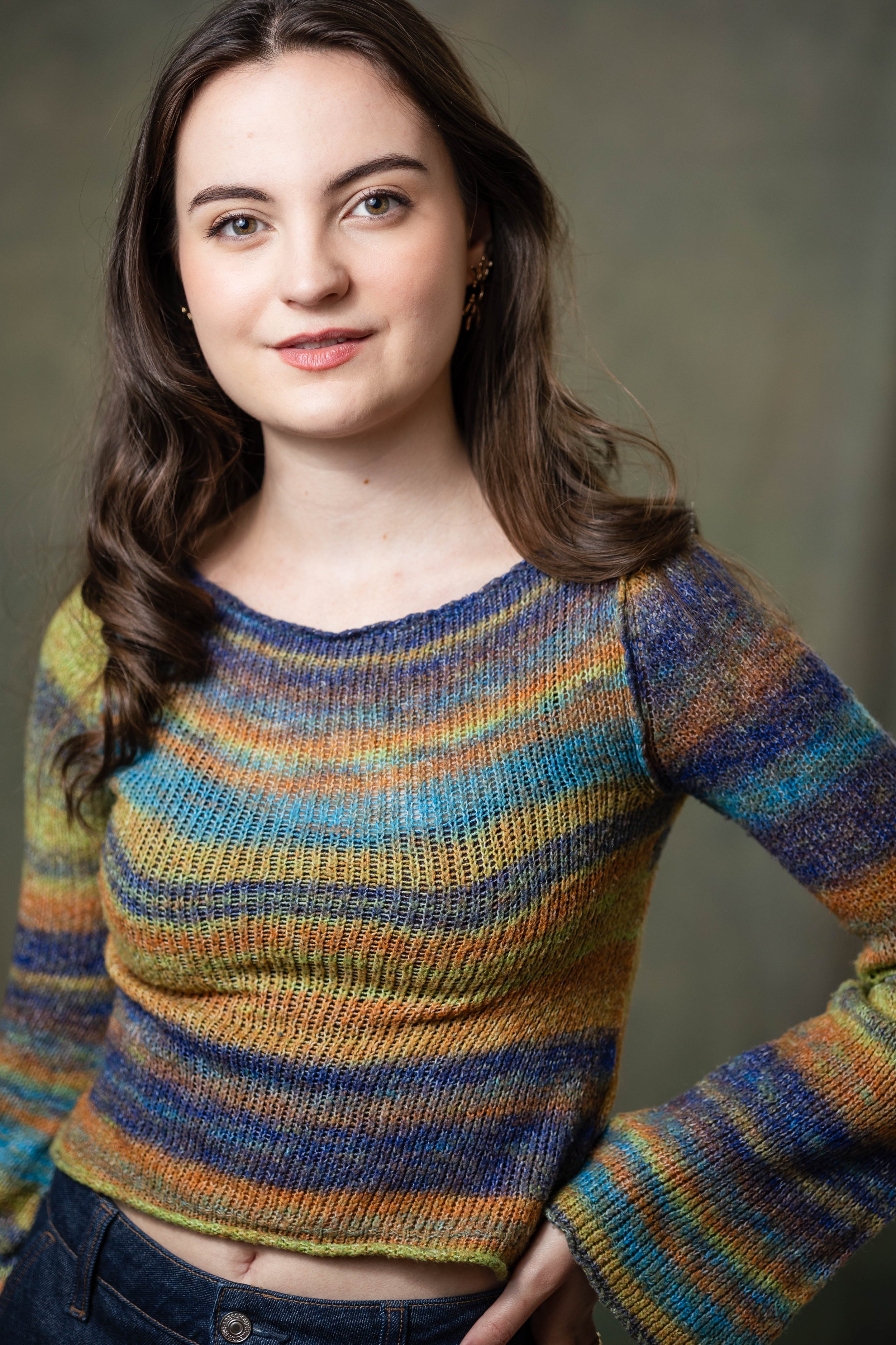 A woman with long wavy brown hair wearing a multicolored striped knit sweater and blue jeans.