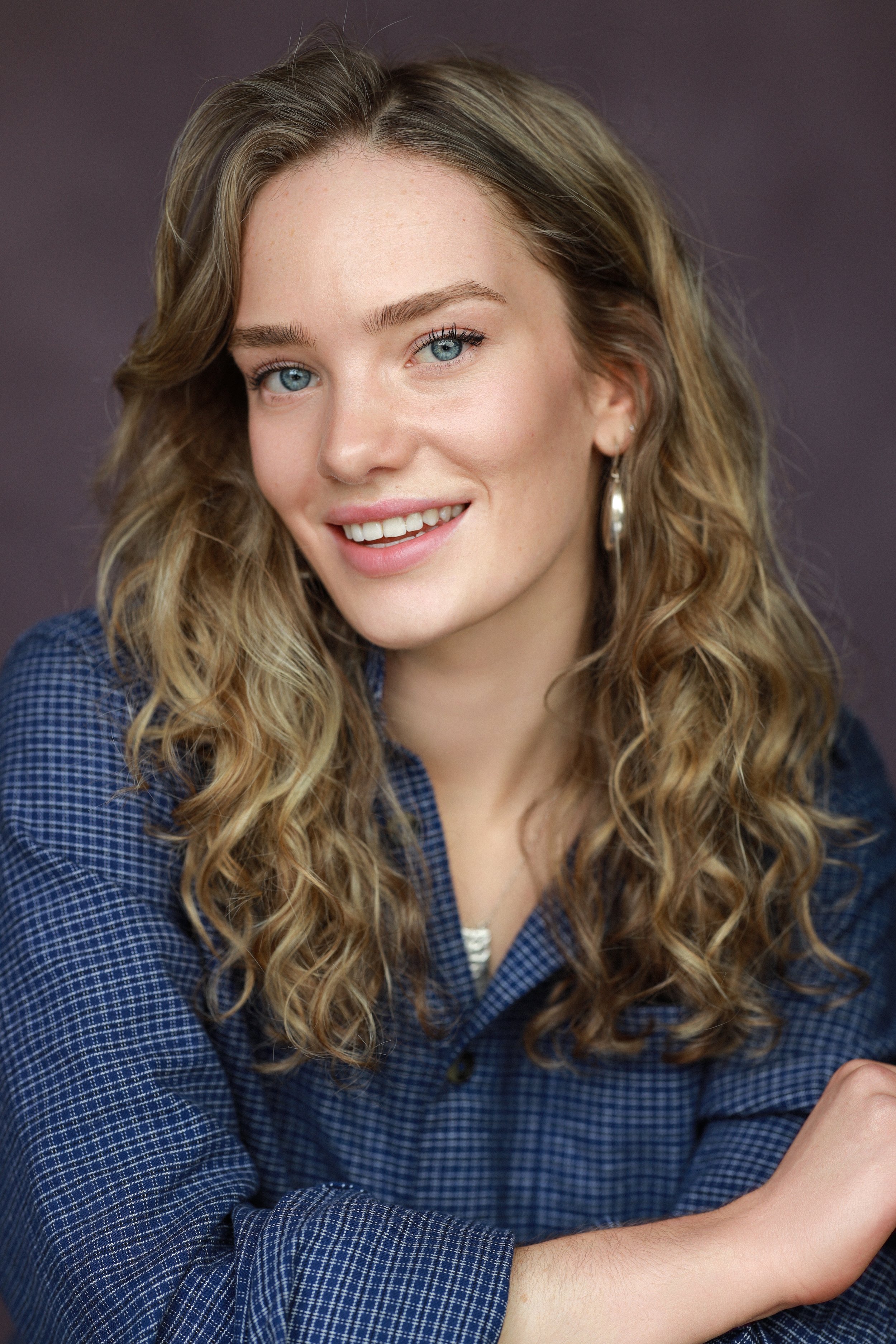 Close-up portrait of a young woman with curly blonde hair and blue eyes, smiling, wearing a blue checkered shirt, against a dark background.