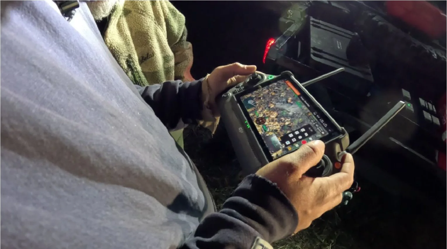 Person using a handheld drone controller with a live video feed on the screen, outdoors at night.