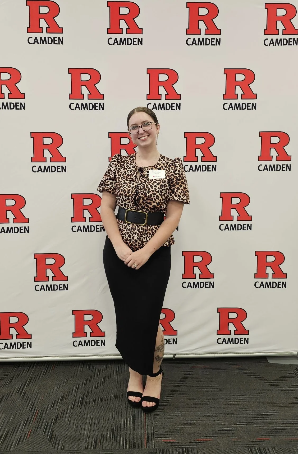 A woman standing in front of a backdrop with red 'R' logos and the word 'Camden' repeated, smiling, wearing a leopard print blouse, black pants, and high heels.