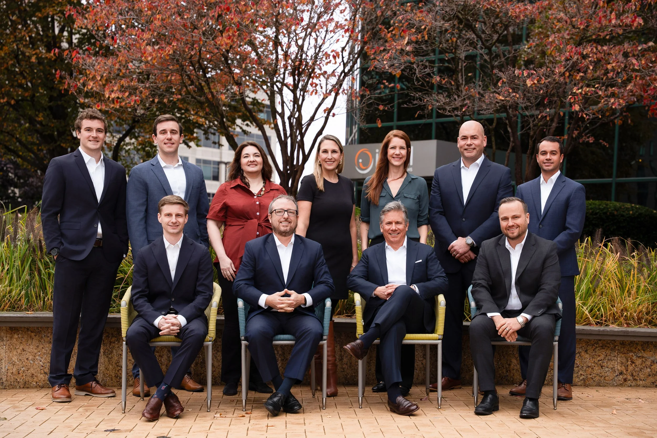 Group of ten diverse professionally dressed people posing outdoors in front of red trees and modern office buildings, sitting and standing on a brick patio.