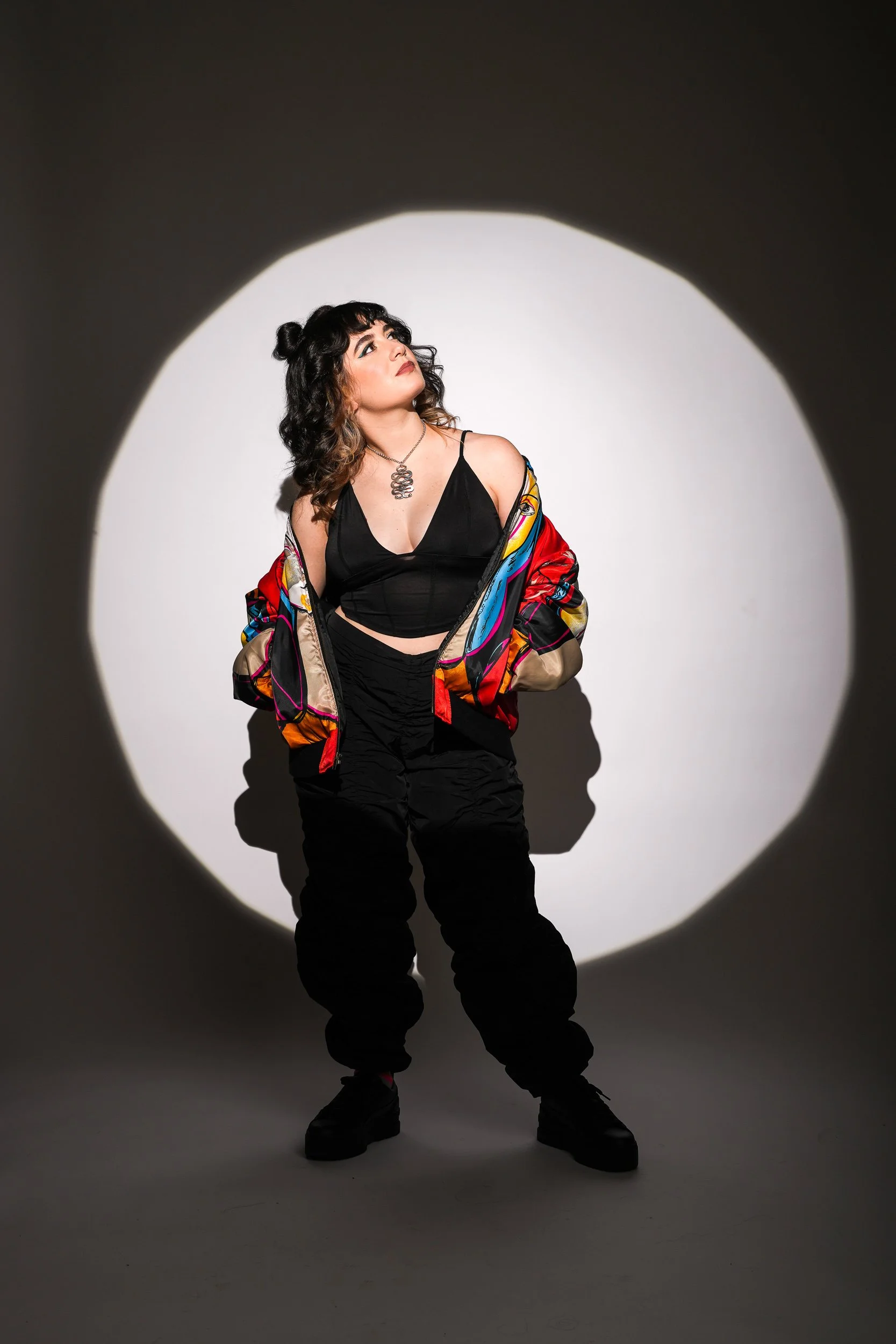 A woman with curly black hair, wearing a black top, colorful jacket, and black pants, stands against a white backdrop with a spotlight creating a circular shadow. She gazes upward, with her hands in her jacket pockets.