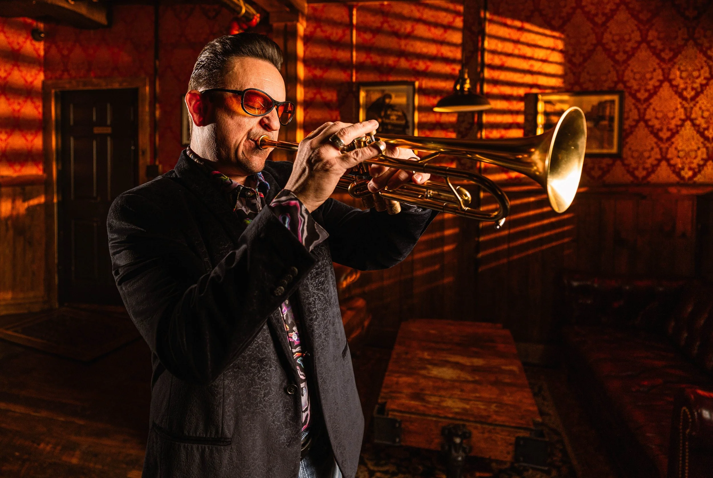 A man wearing sunglasses and a black jacket playing a trumpet in a dimly lit room with wooden walls and framed pictures.