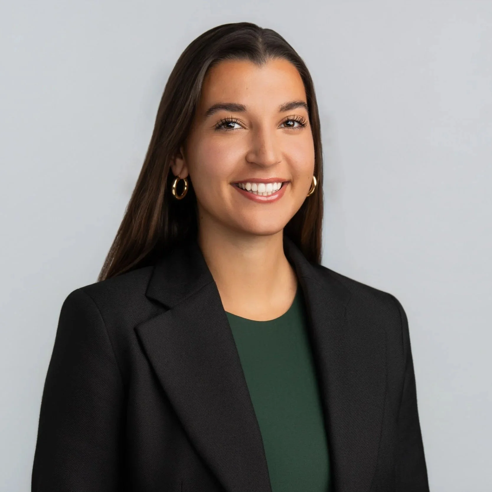 A woman with long dark hair wearing a black blazer, green top, and gold hoop earrings, smiling against a plain light gray background.