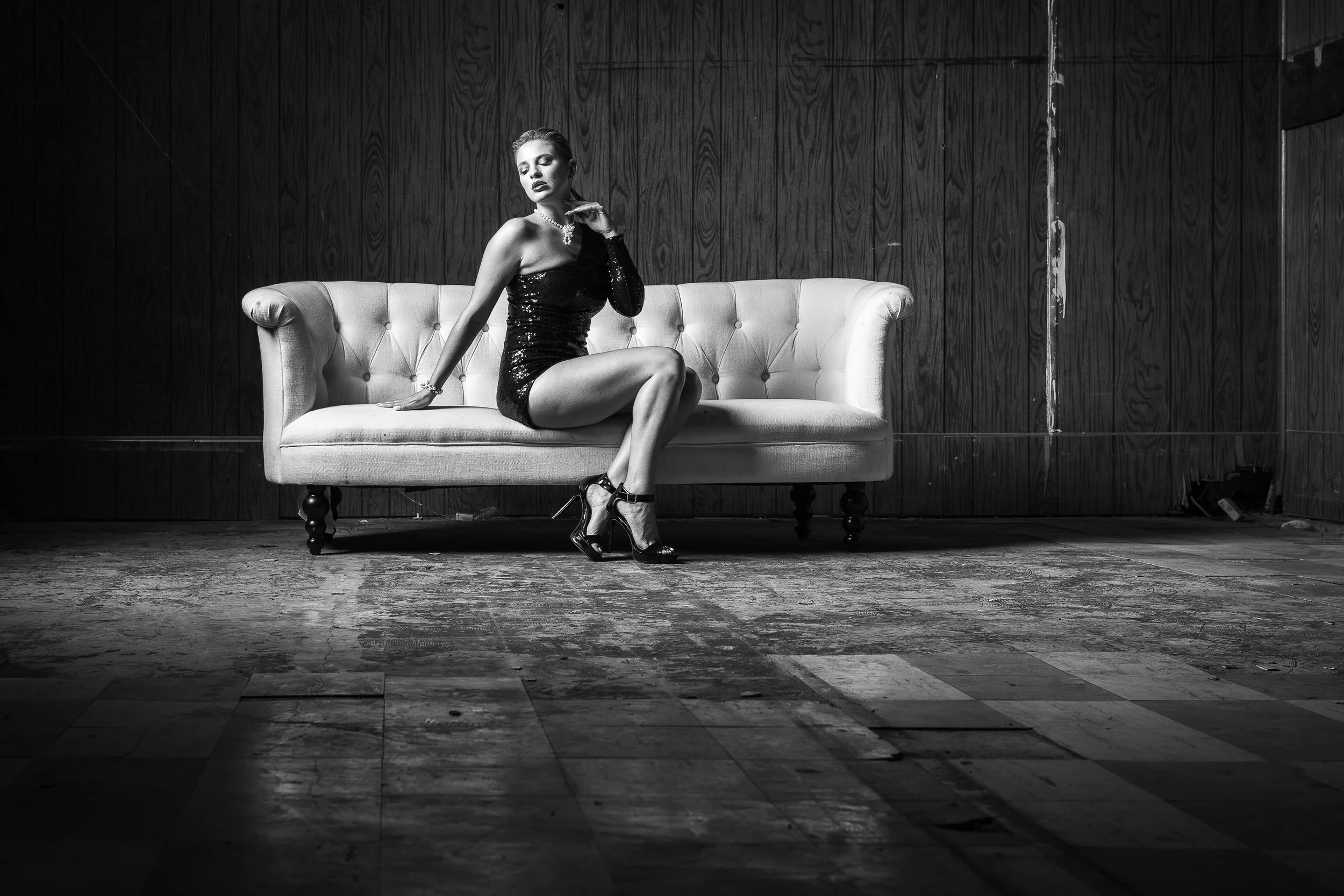 A woman in a black sequined dress and high heels sitting on a vintage sofa with a pearl necklace, posed in front of a dark wooden wall in a black and white photograph.