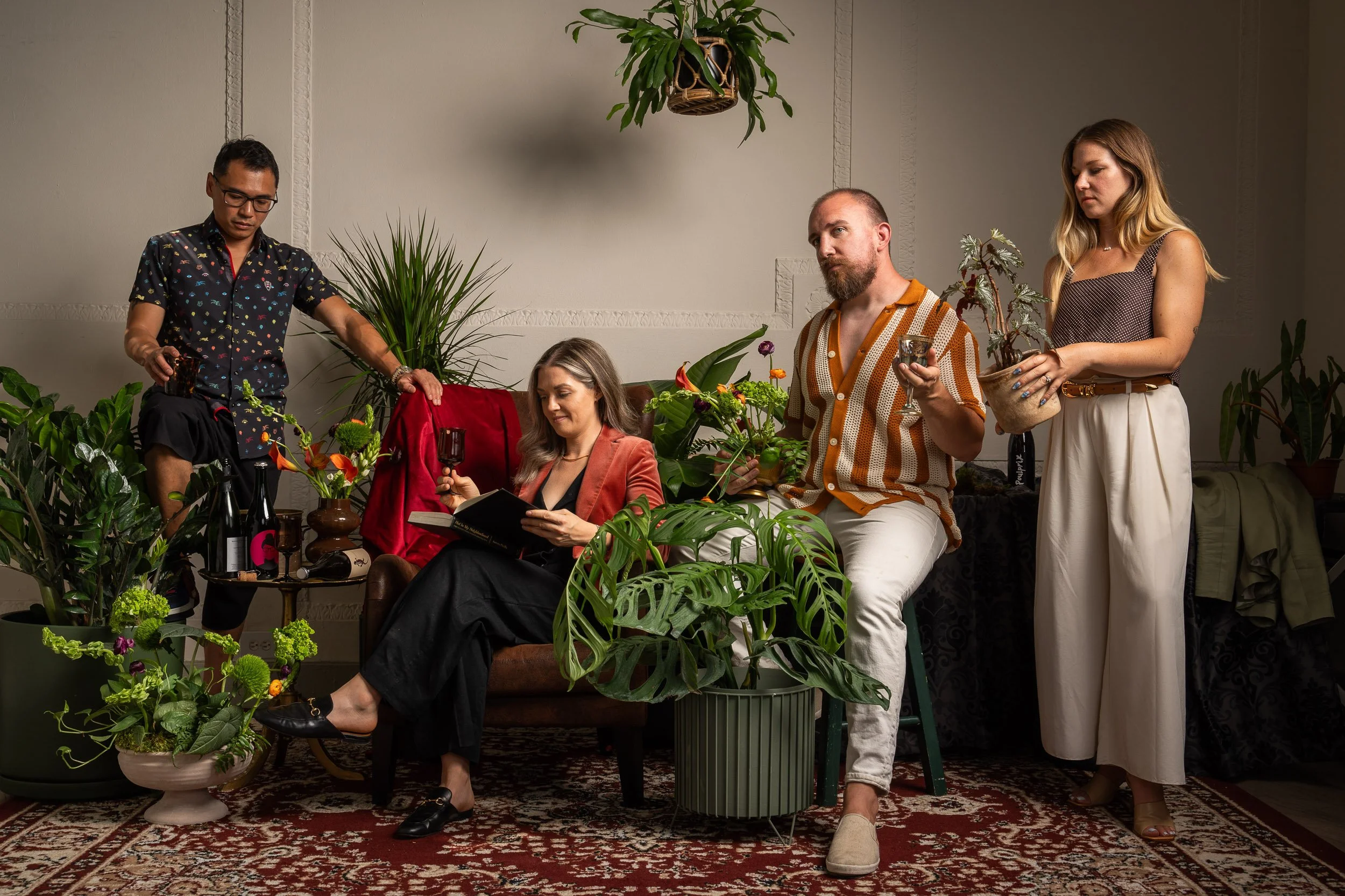A group of five people in an indoor space with plants. One woman is sitting on a chair reading a book while holding a glass of wine. Two women and one man are standing around her, with one woman holding a potted plant and a glass of wine, one woman holding a potted plant, and the man holding a glass of wine. The space has a carpeted floor and decorative wall trim.