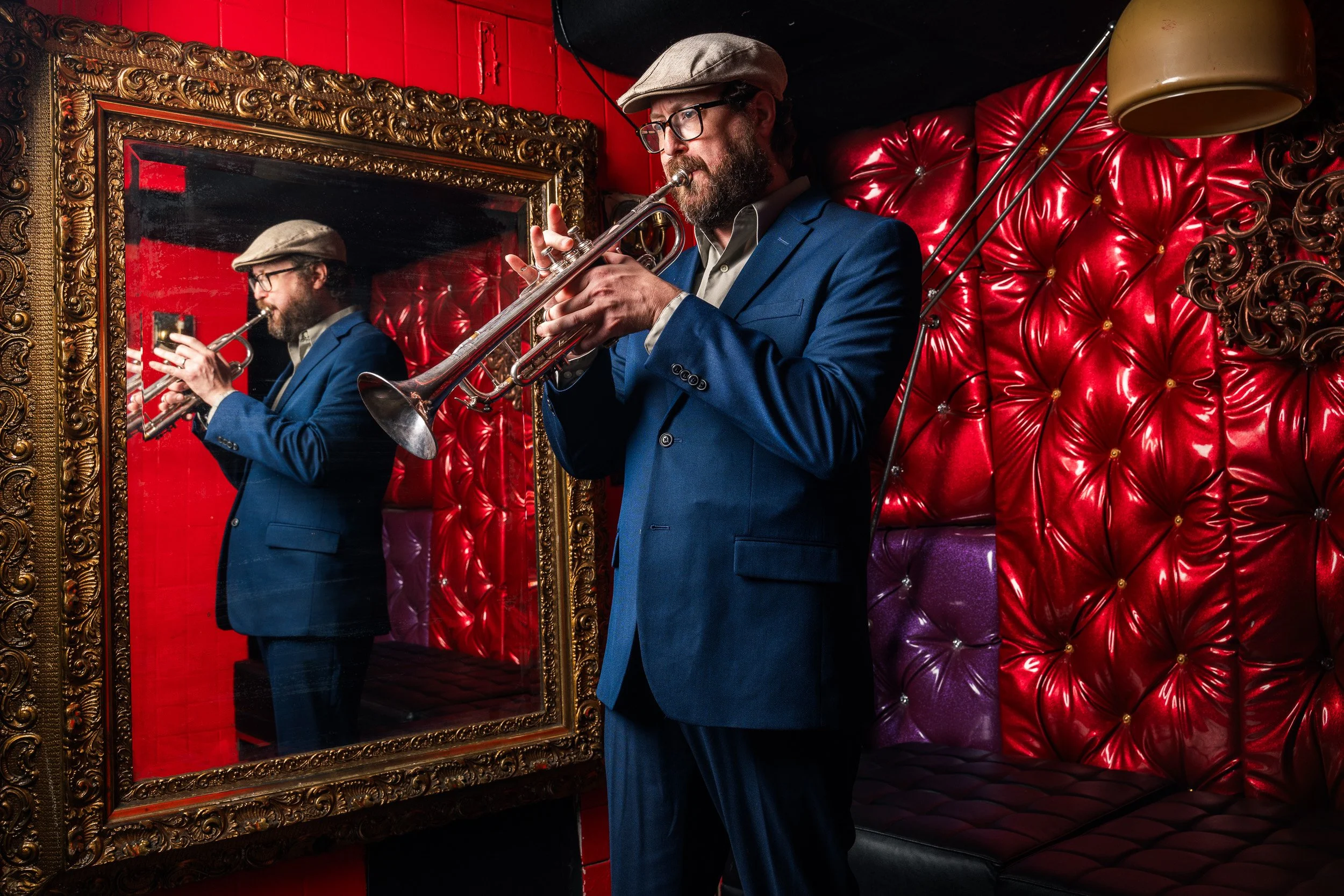 A man with glasses, a beard, and a flat cap playing a trumpet in a room with ornate gold-framed mirror and red velvet tufted wall.