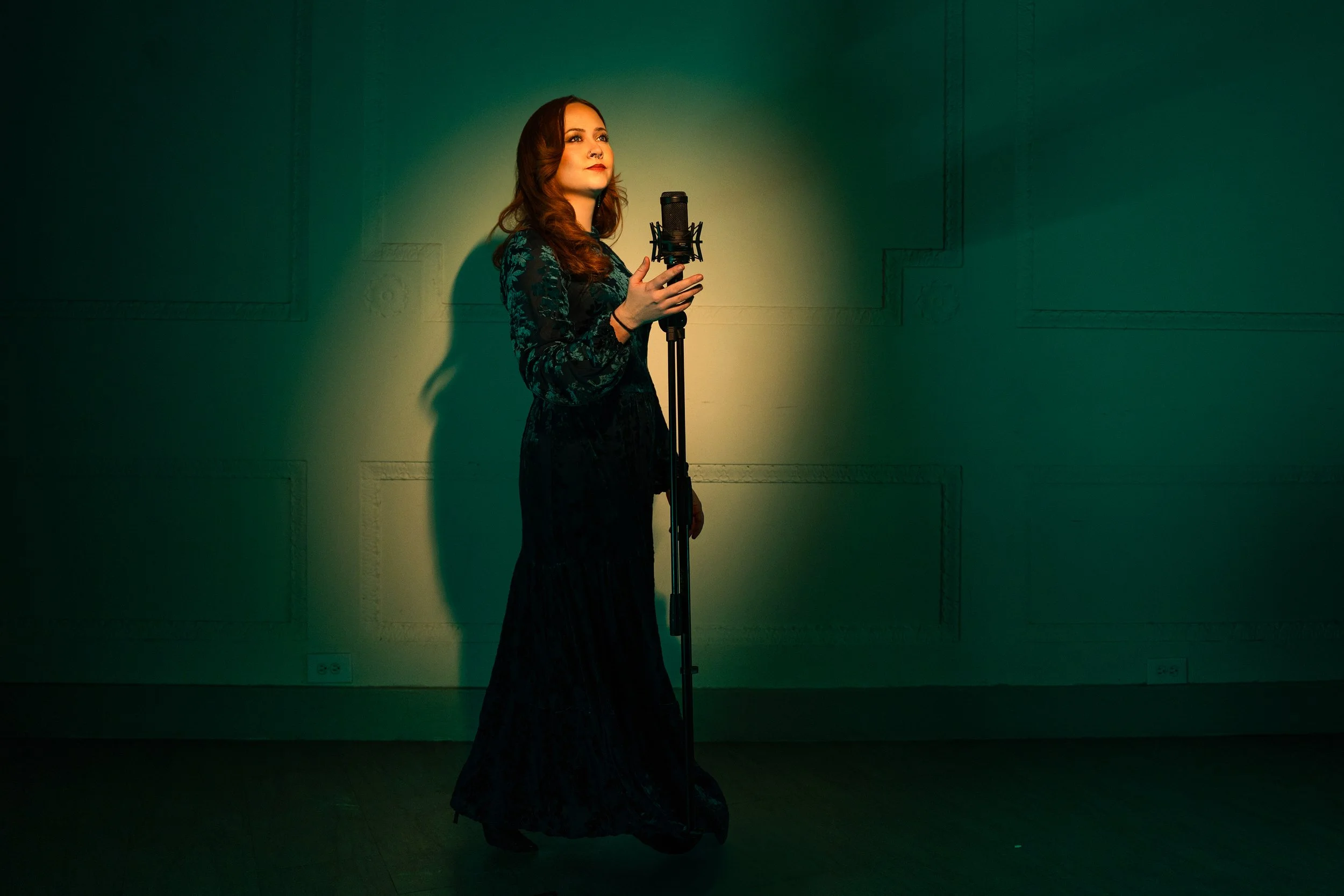 A woman with long brown hair in a dark floral dress stands in front of a green wall, holding a microphone and looking slightly upwards, illuminated by soft yellow lighting.