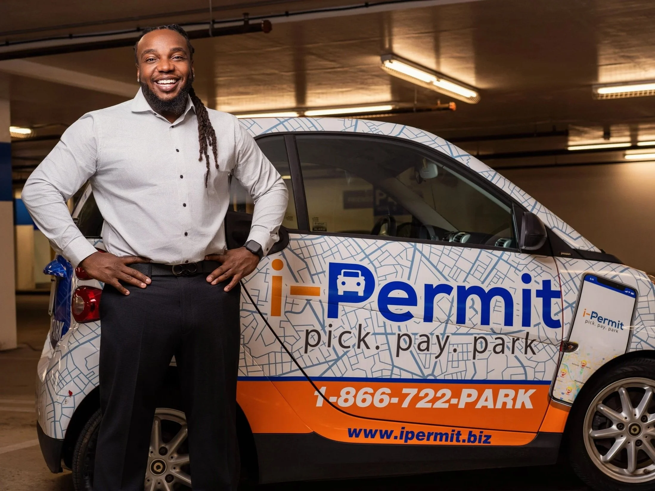 A smiling man with long dreadlocks wearing a light gray button-up shirt and dark pants standing in front of an electric vehicle with branding for iPermit, a parking permit service, inside a parking garage.