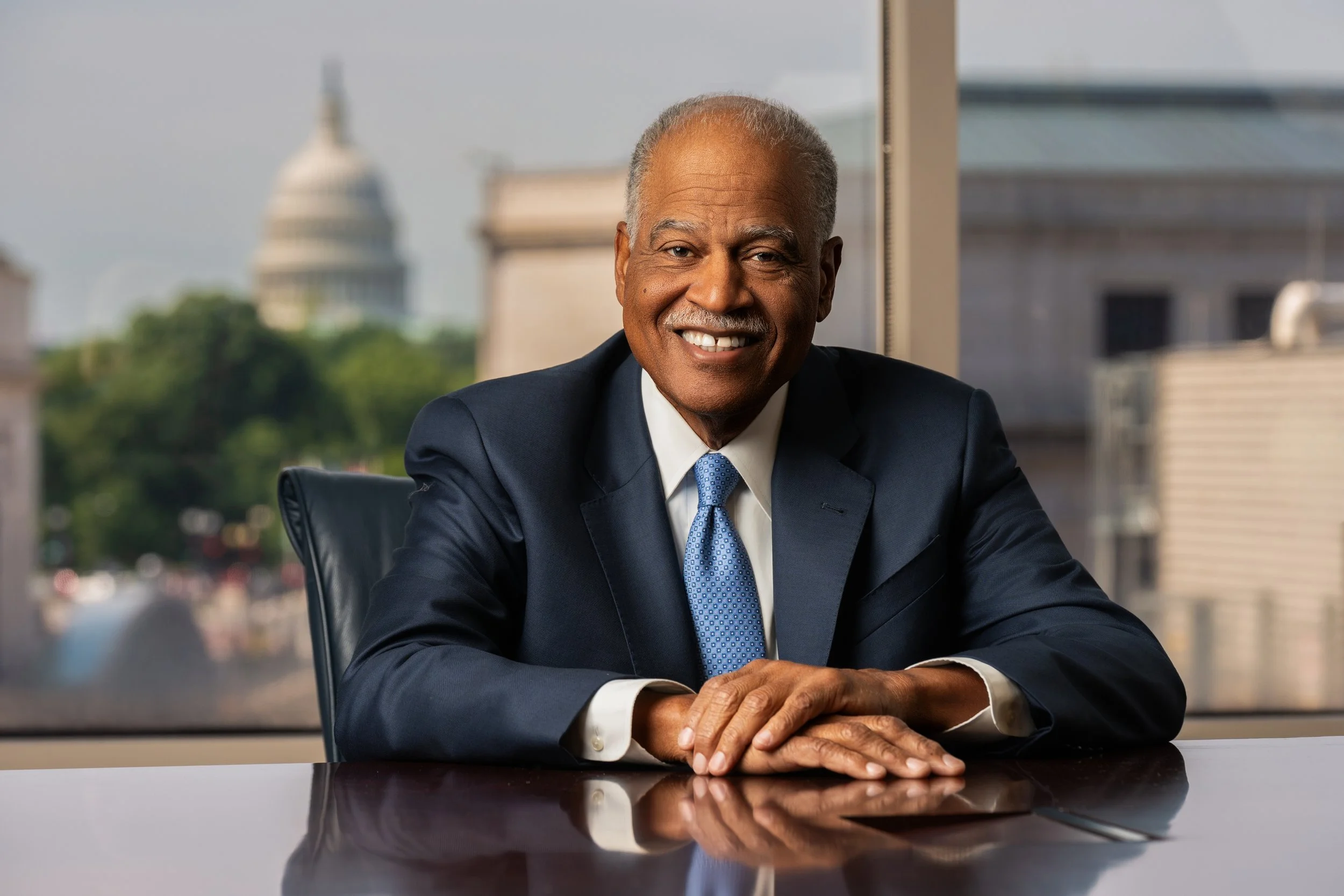 smiling elderly man in a suit sitting at a dark wood conference table with arms crossed, window behind showing blurred buildings and Capitol dome in the background