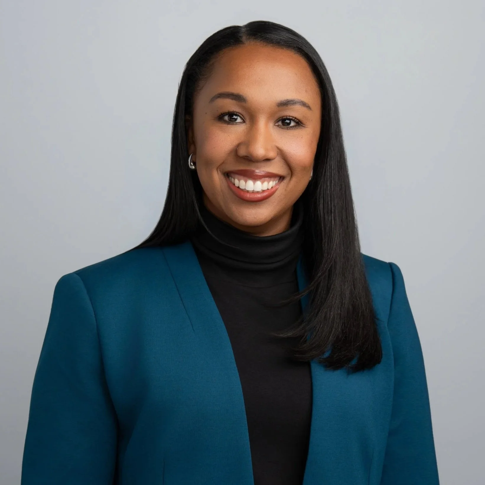 A woman with dark hair, wearing earrings, a black turtleneck, and a blue blazer, smiling against a plain light gray background.