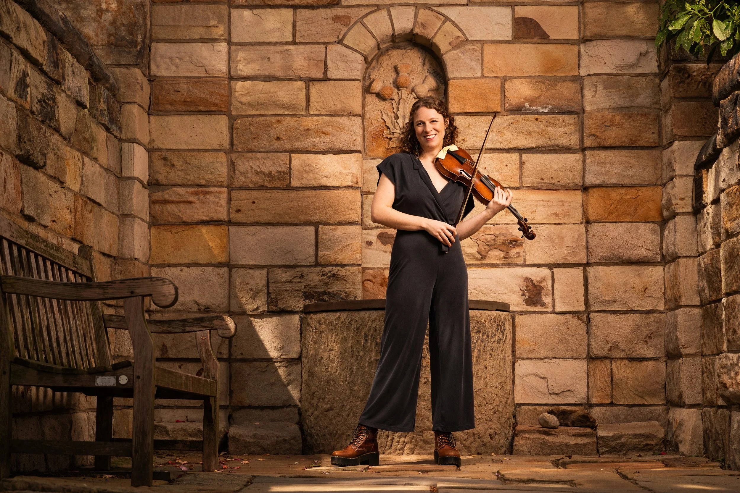 A woman standing outdoors in front of a stone wall, holding a violin and smiling, wearing a dark jumpsuit and lace-up boots.