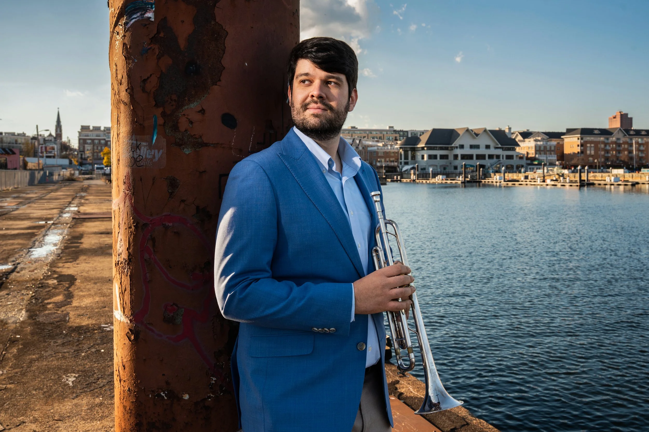 A man in a blue suit holding a trumpet, standing by a waterway with boats in the background, next to a weathered, graffiti-covered pole.