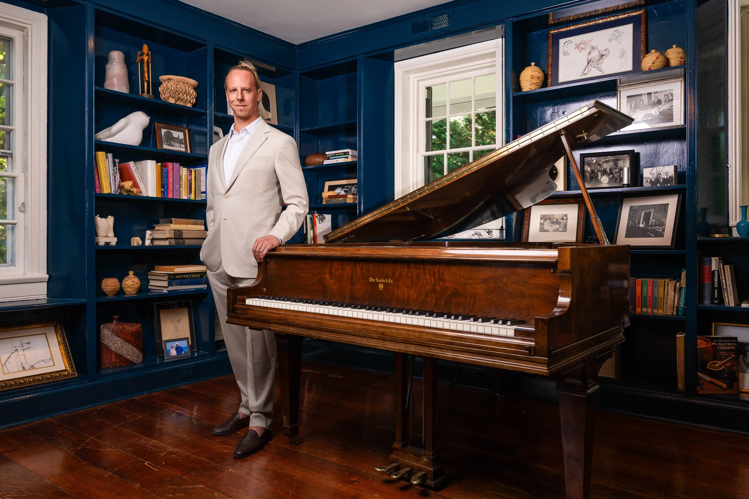 A man in a beige suit standing next to a grand piano in a room with blue shelves filled with books, framed photographs, and art pieces, with a window showing outdoor greenery.