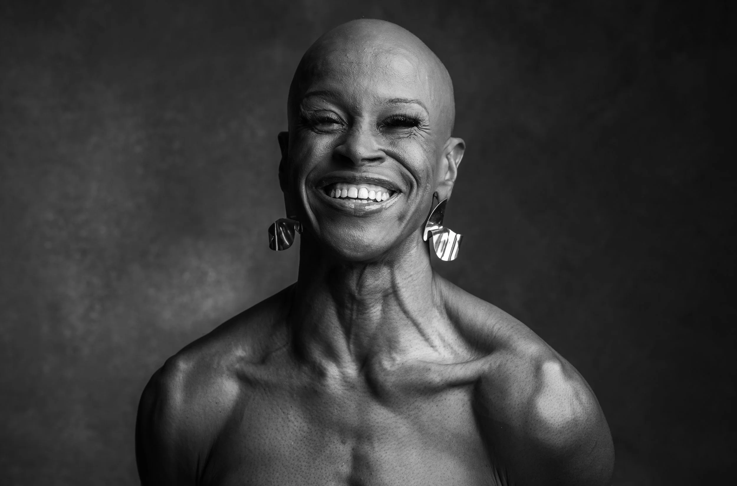 Black and white portrait of a smiling woman with a bald head, wearing large earrings, bare shoulders, and closed eyes.