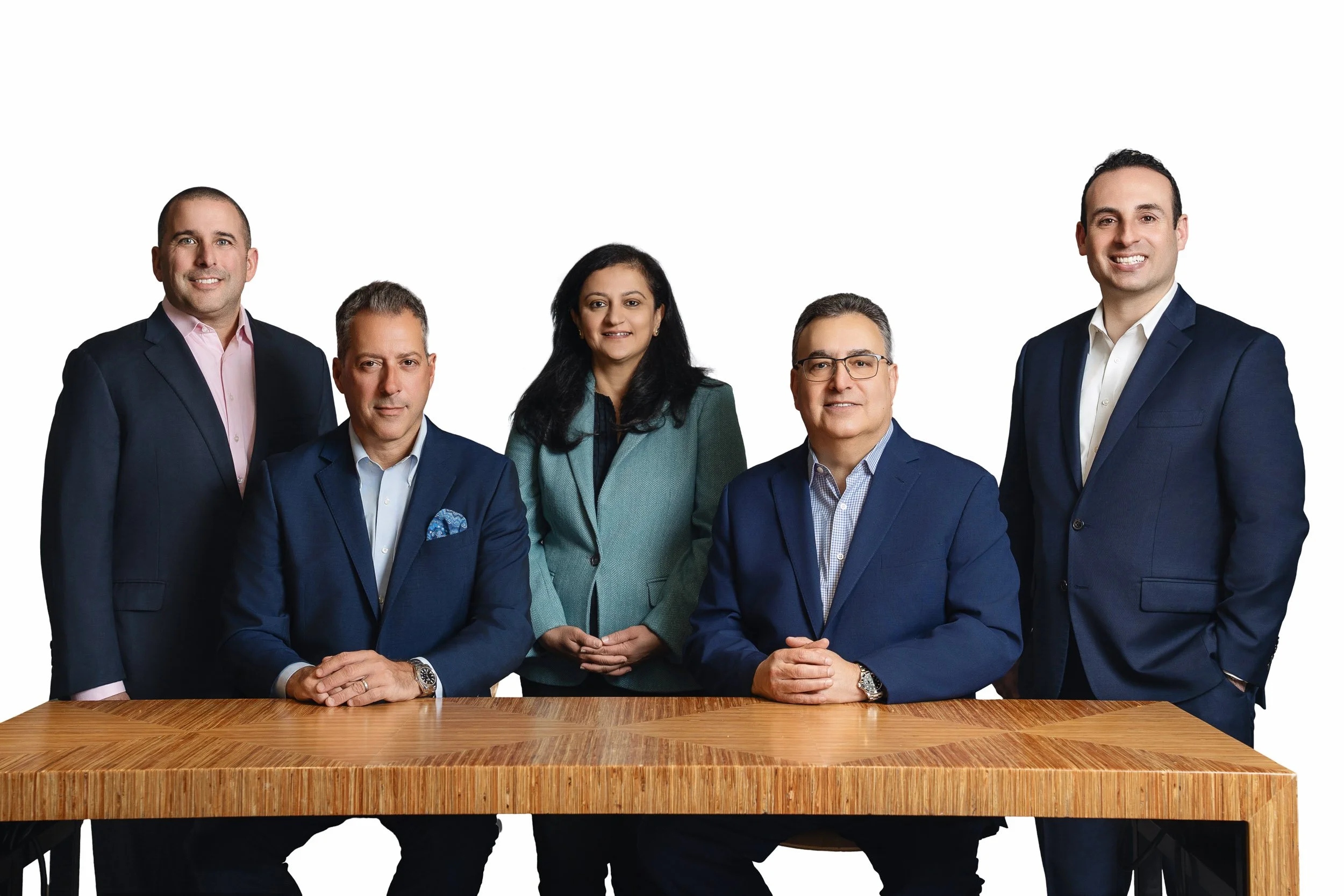 Group of six professionally dressed people, five men and one woman, standing and sitting behind a large wooden table, smiling at the camera, against a plain white background.