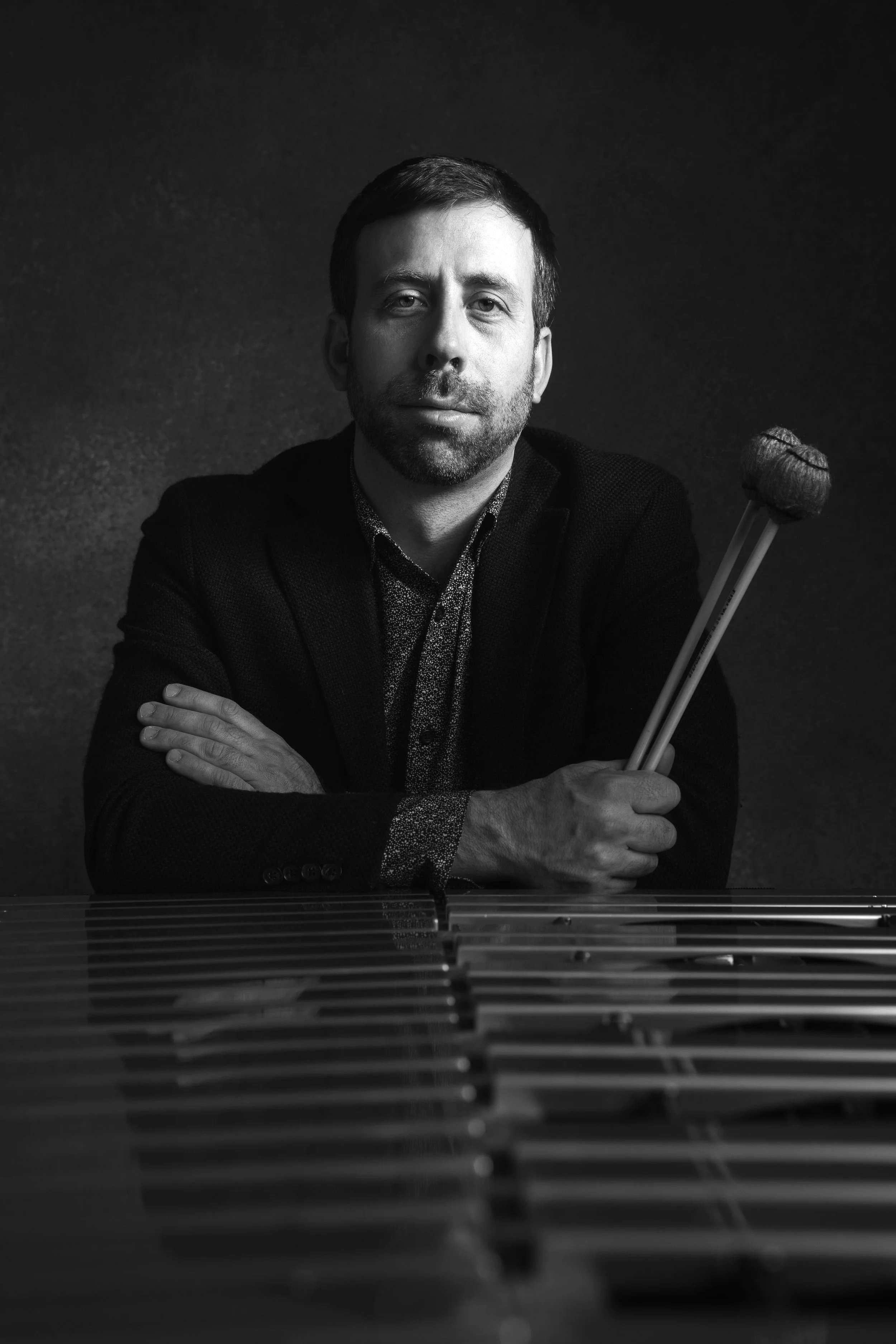 Black and white portrait of a man with short hair and a beard, sitting with crossed arms. He has a calm expression and is holding drumsticks, with a percussion instrument in front of him. The background is dark.